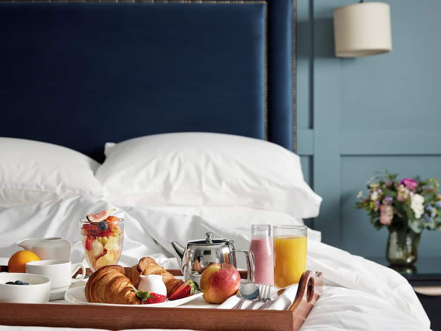 Una bandeja de desayuno bien dispuesta sobre la cama, con diversos alimentos, una tetera y un vaso de zumo de naranja.