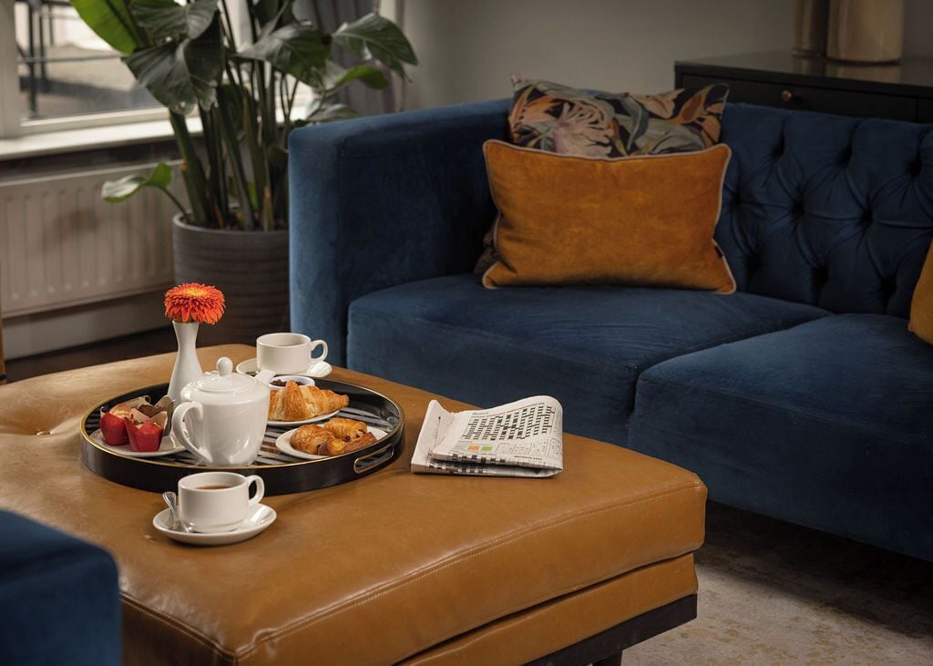 A cozy living room with a blue sofa, a brown ottoman, and a tray with breakfast items including croissants, a teapot, cups, and a newspaper.