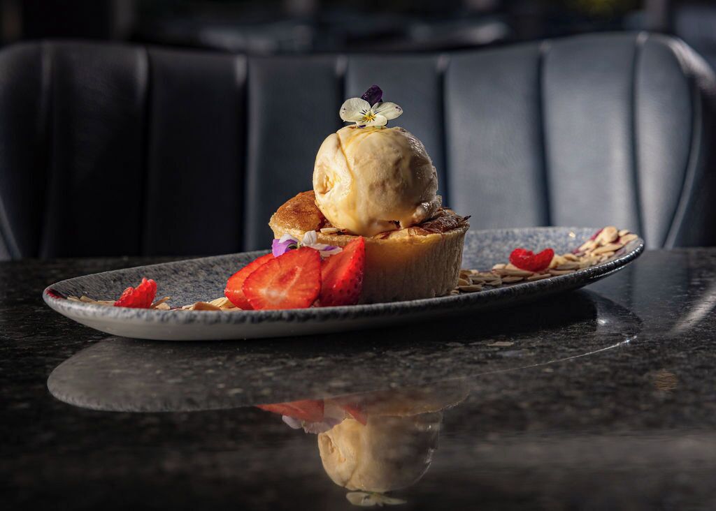 Un dessert présenté sur une assiette, surmonté d’une fleur, accompagné de fraises, et dont le reflet se voit sur la table.