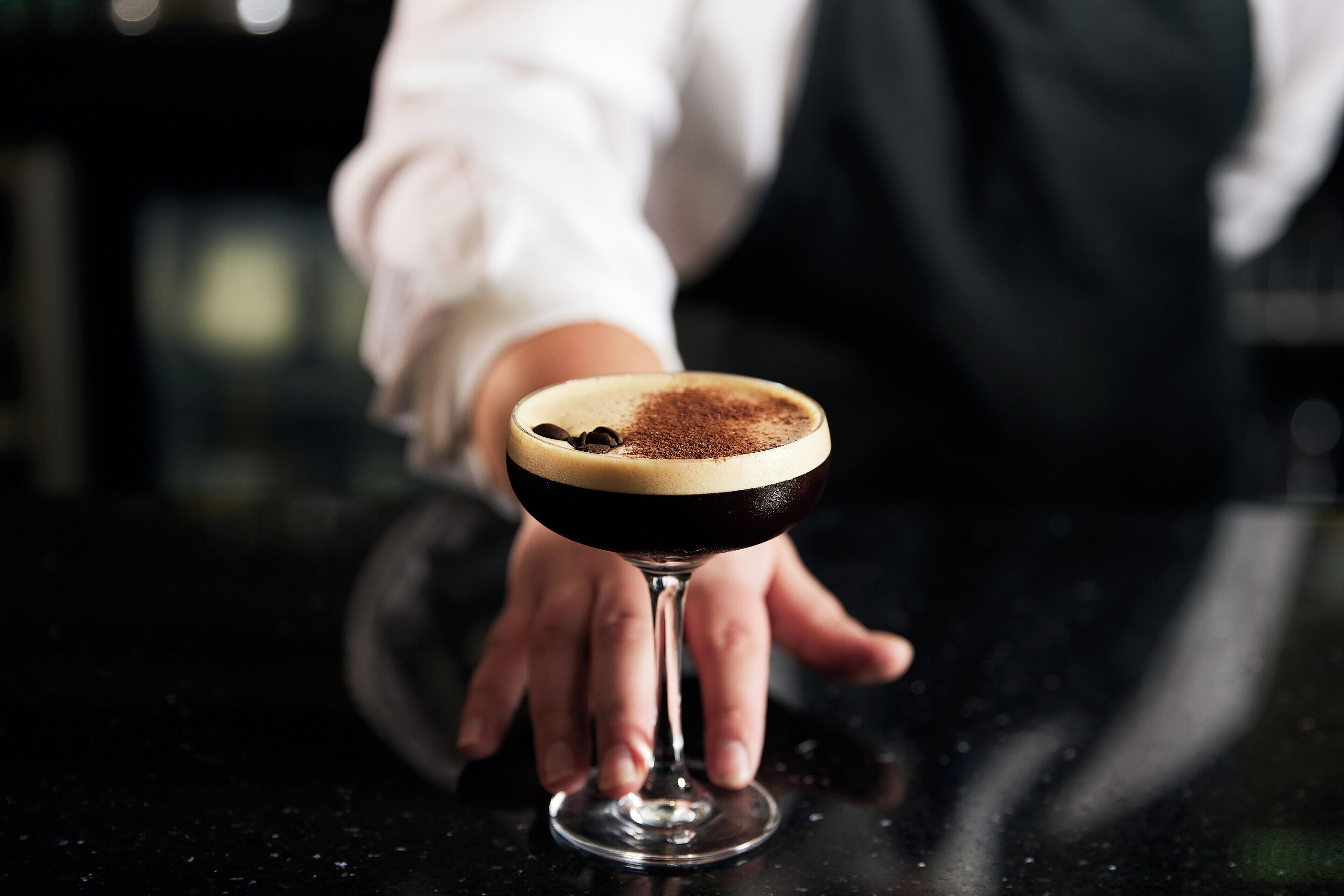 Ein Cocktail im Stielglas mit dunkler Flüssigkeit und hellem Schaum, auf einer reflektierenden Oberfläche platziert.