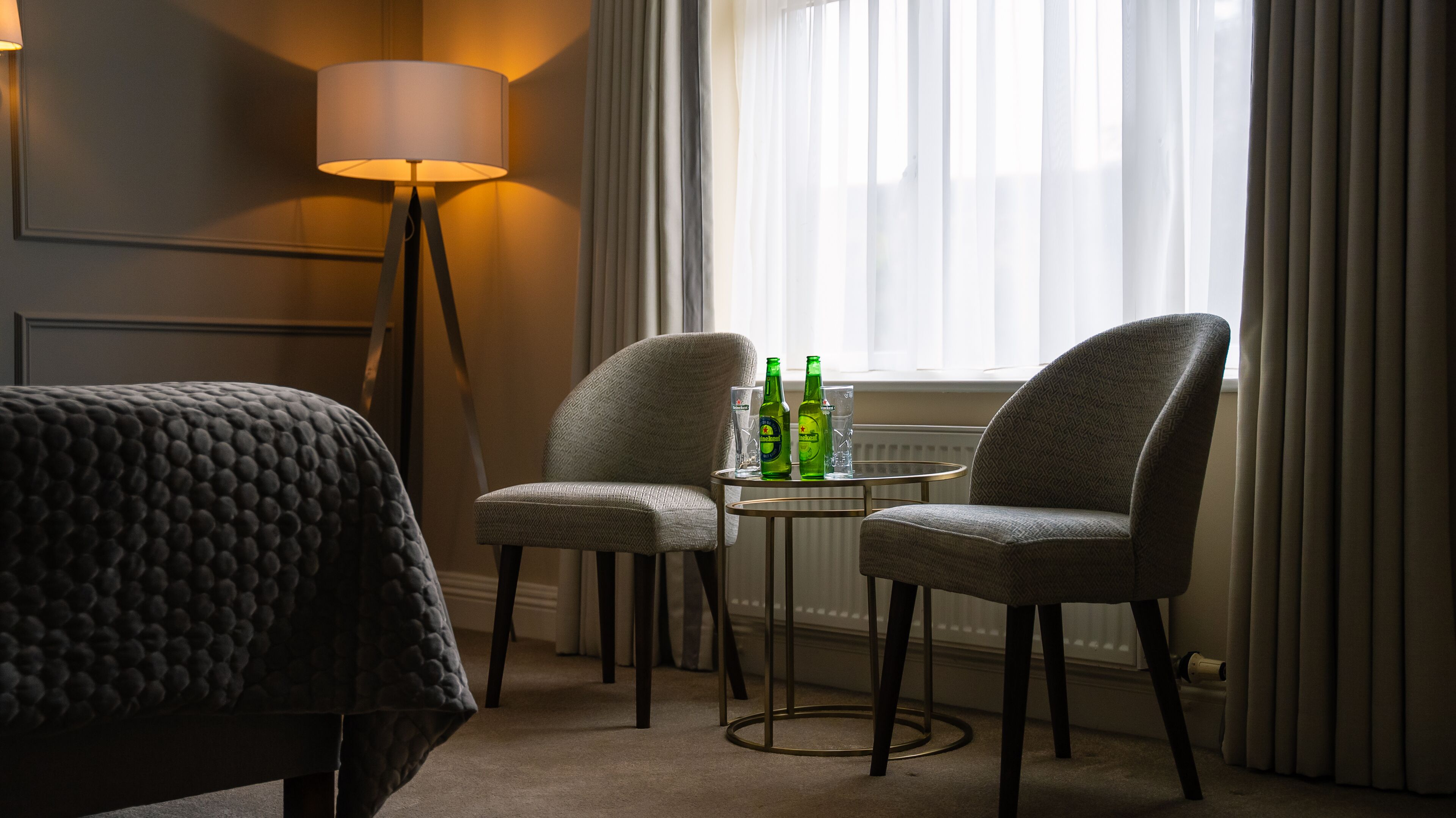 A hotel room with a bed, two armchairs, a small table with bottles and glasses, a lamp, and a window with curtains.
