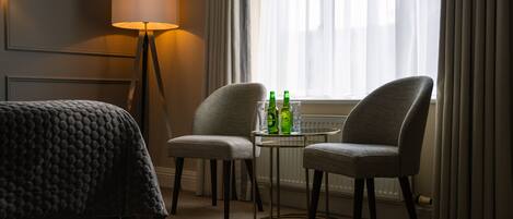 A hotel room with a bed, two armchairs, a small table with bottles and glasses, a lamp, and a window with curtains.