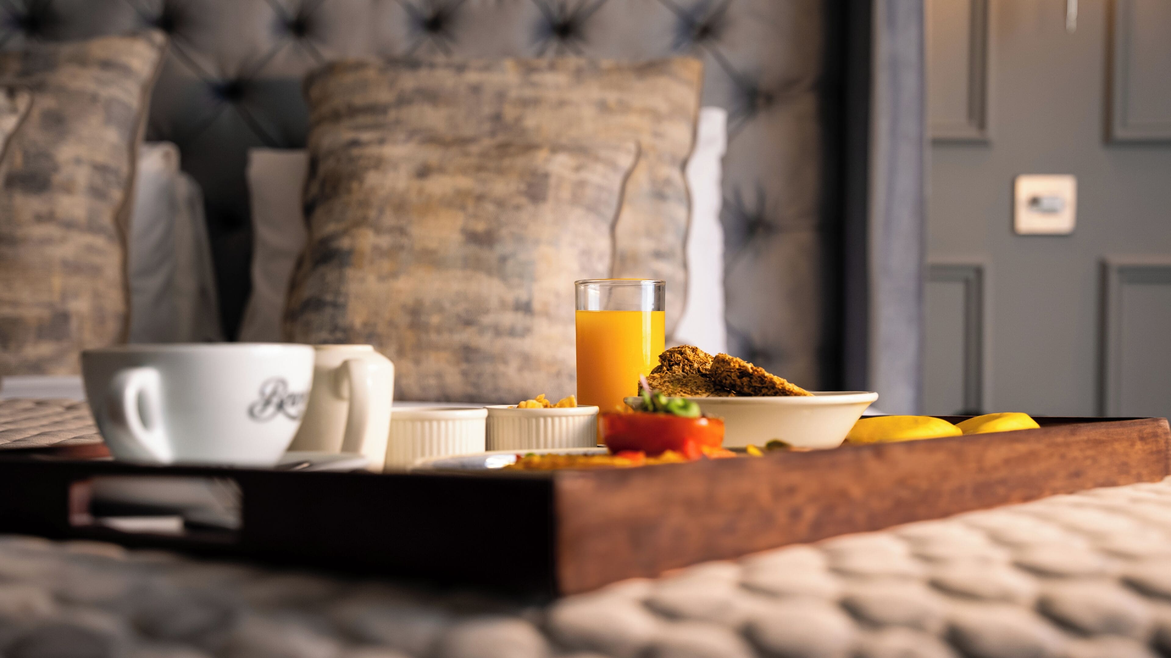 Bandeja de desayuno con taza, vaso de zumo de naranja y bol de cereales sobre una cama con cabecero capitonado.