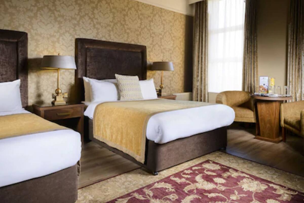 A hotel room with two beds, a large wooden headboard, a red patterned rug, and a small side table with a lamp.