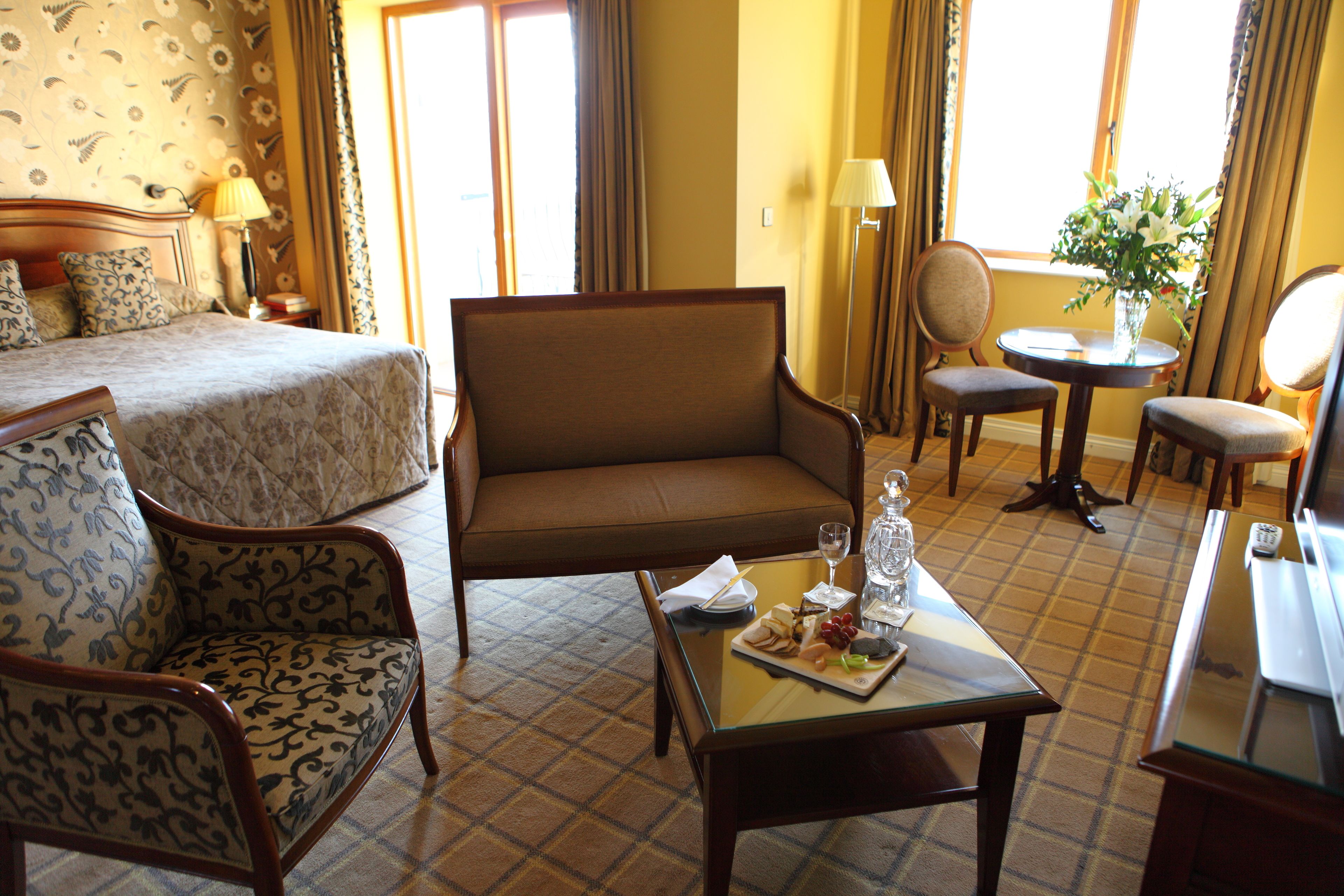 A hotel room with a bed, a sofa, a dining table with a glass of wine, and a vase with flowers.