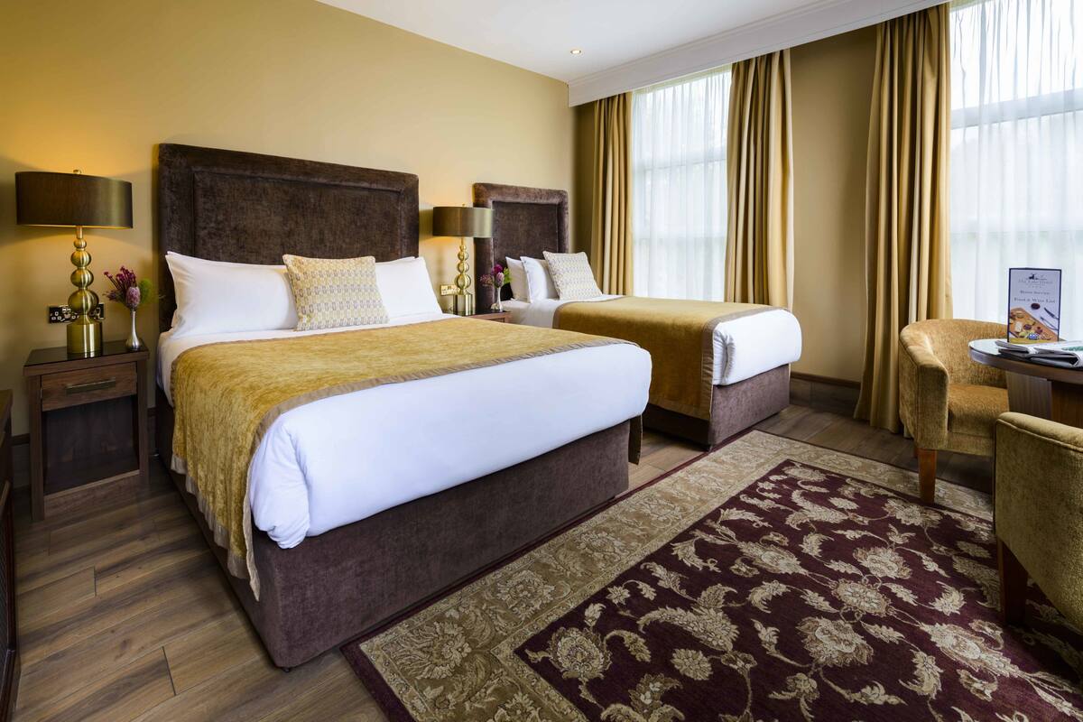 A hotel room with two beds, a large window with curtains, a desk with a chair, and a decorative rug.
