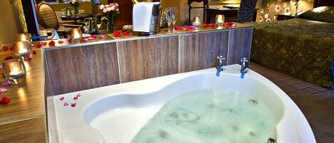 Eine große Badewanne voller Wasser, umgeben von Rosenblüten, in einem Raum mit Holzboden und einer gemusterten Wand.
