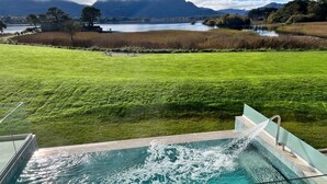 Una piscina con acqua che si riversa sul bordo, un prato verde e un lago con montagne sullo sfondo.