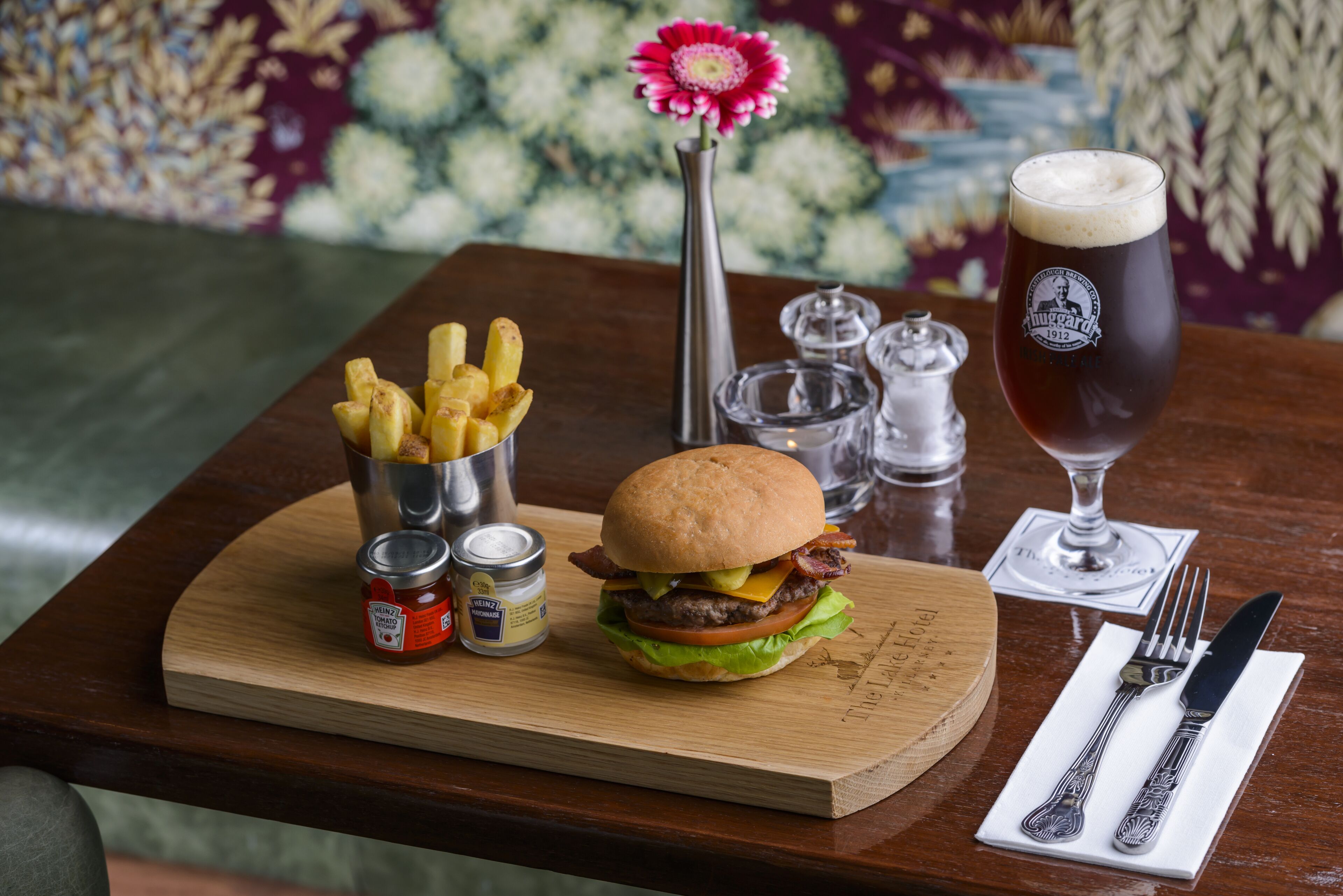 Uma refeição com um hambúrguer, batatas fritas e uma cerveja em uma tábua de madeira.
