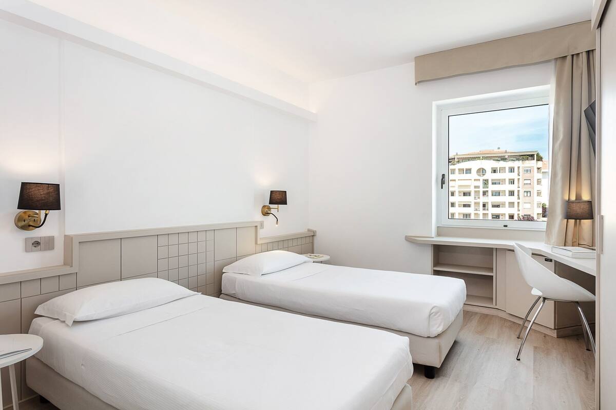 A hotel room with two beds, a desk, a chair, a window with a view of buildings, and wall-mounted lamps.