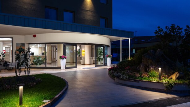 A hotel entrance with a curved facade, glass doors, and a landscaped garden area with potted plants and a pathway. - Hotel Carlo Felice (Sassari)