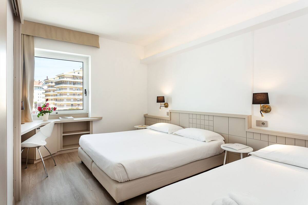 A hotel room with two beds, a desk, and a window with a view of other buildings.