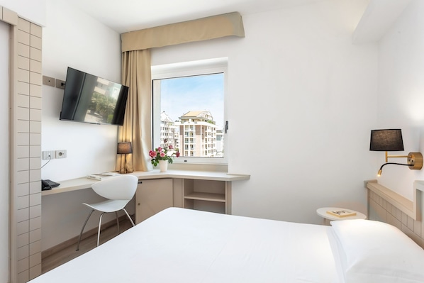 A hotel room with a bed, a desk with a chair, a TV, a lamp, and a view of buildings through the window.