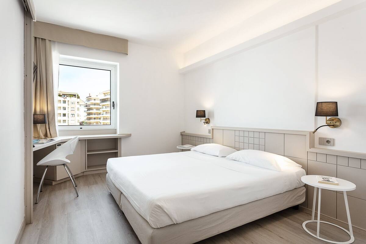 A hotel room with a bed, a desk, and a chair. There is a window with curtains, and two wall-mounted lamps.