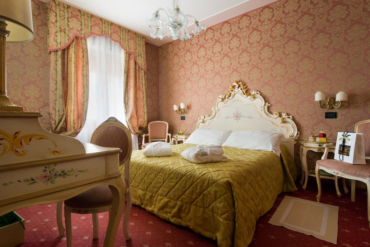 A hotel room with a bed, a desk, a chair, a small table, and a chandelier.