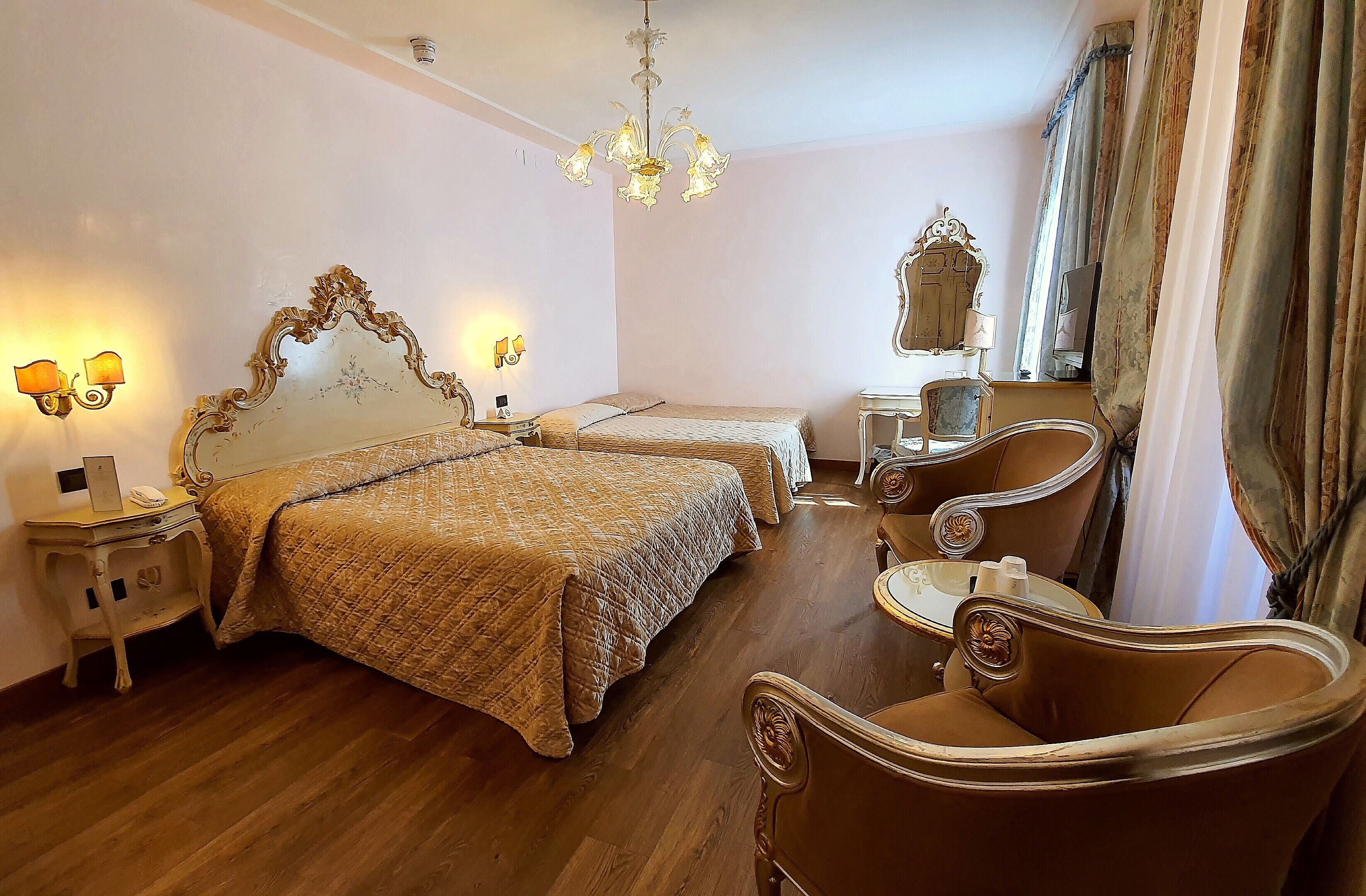 Ein Hotelzimmer mit einem großen Bett, einem kleineren Bett, einem Schminktisch mit Spiegel, einem Sessel und einer Chandelier.