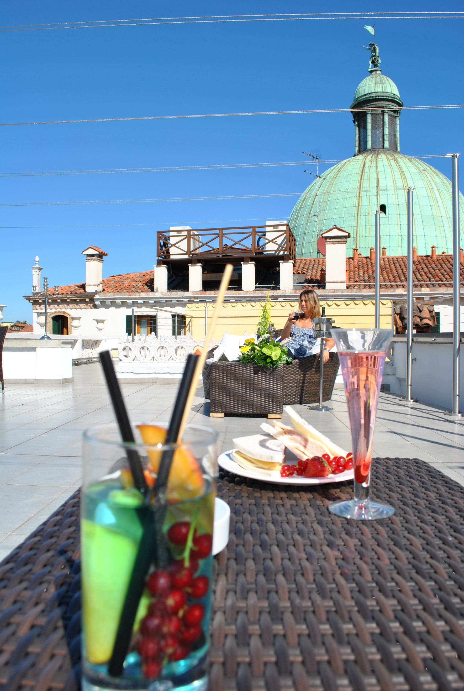Una mesa con bebidas y un plato de comida, un edificio con cúpula al fondo, y un cielo azul despejado.