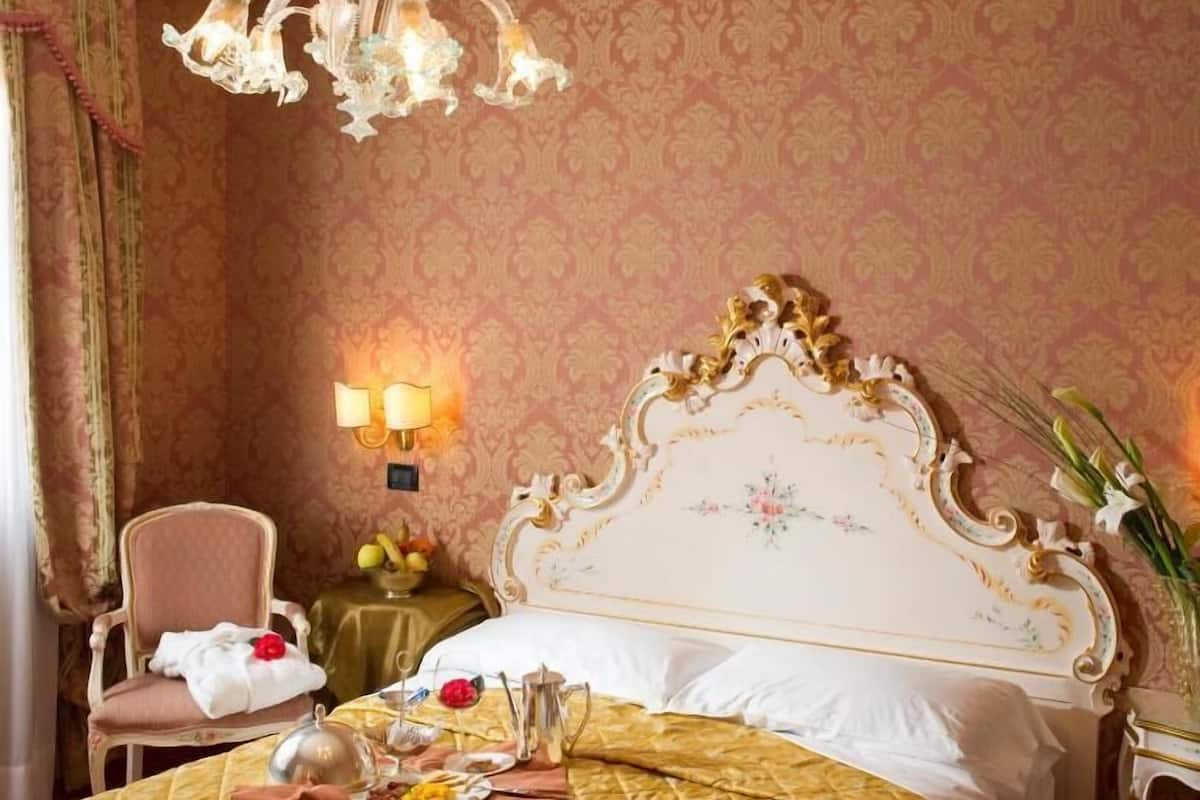 A hotel room with a large bed, a chandelier, a chair, a table with a vase of flowers, and a wall with patterned wallpaper.