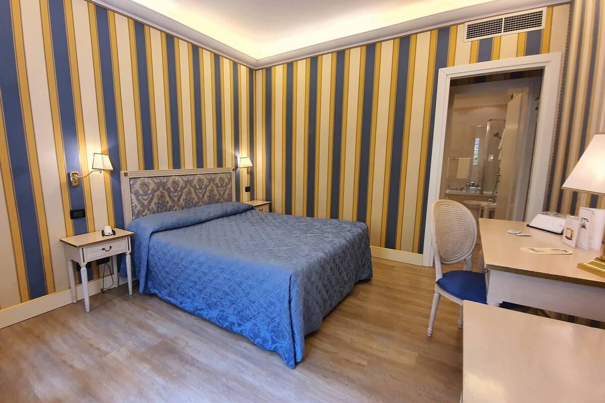 A hotel room with striped wallpaper, a bed with a blue bedspread, a bedside table with a lamp, and a desk with a chair.