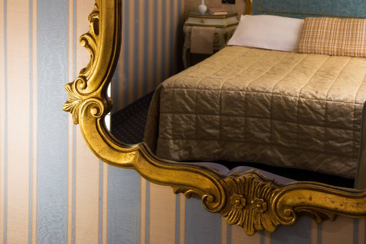 A room with a large ornate mirror, a bed with a quilted cover, a nightstand with a lamp, and striped wallpaper.