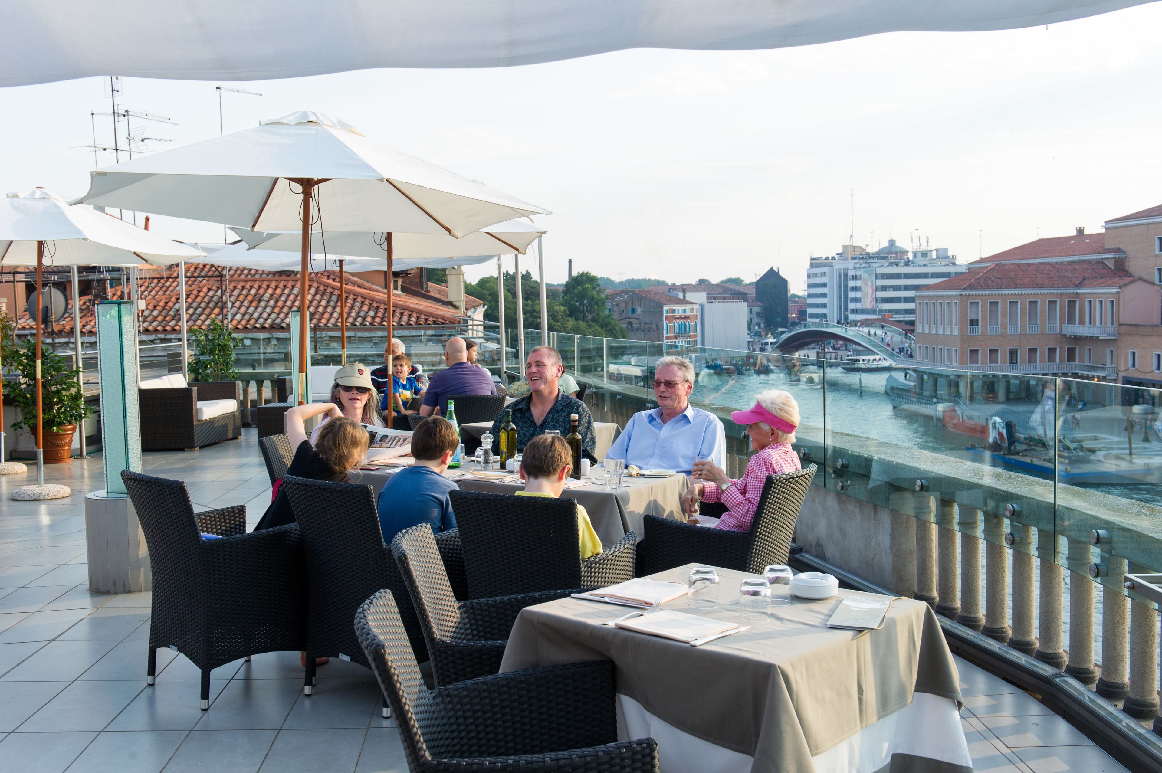 A group of people dining at an outdoor table with a view of a canal and buildings in the background.