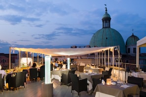 An outdoor dining area with tables and chairs, a large tent, and a view of a domed building in the background. - Hotel Carlton on the Grand Canal (Venice)