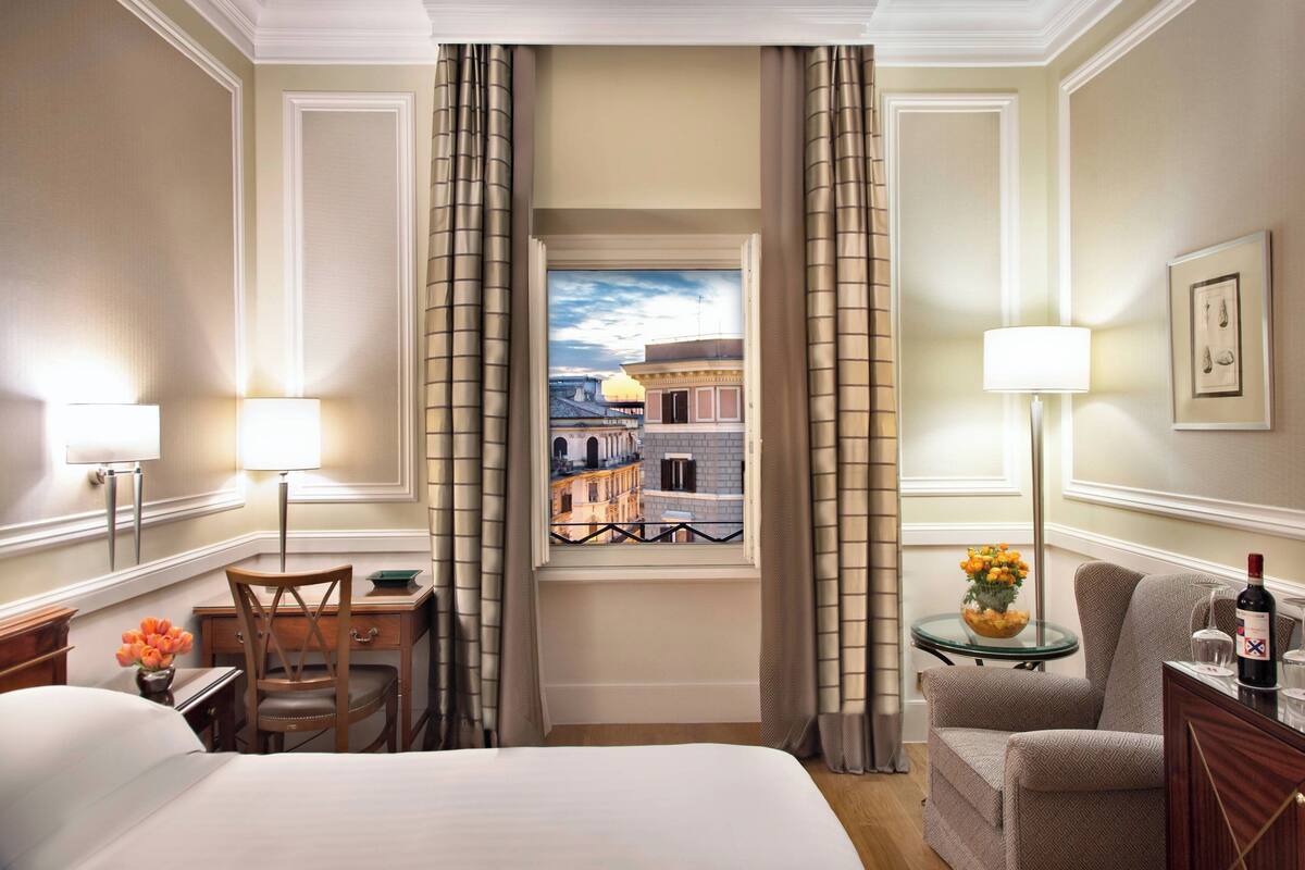 A hotel room with a bed, a desk with a chair, a small table with a lamp, a chair, and a view of buildings through a window with curtains.