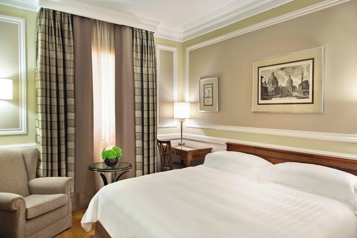 A hotel room with a large bed, a chair, a small table with a lamp, and a framed picture on the wall.