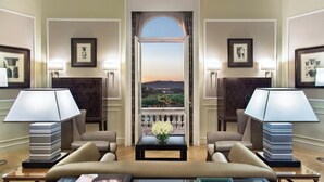 Une chambre d’hôtel avec un grand lit, deux fauteuils, une table basse et une vue sur le paysage urbain, visible à travers une fenêtre en arc de cercle.
