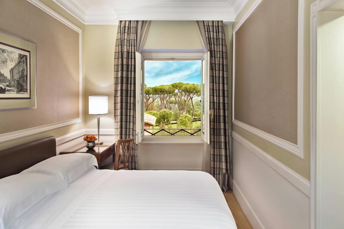 A hotel room with a bed, a nightstand with a lamp, a framed picture on the wall, and a window with curtains.