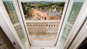 Une vue depuis un balcon, offrant une perspective dégagée sur un bâtiment historique doté d’arcades et d’un toit rouge, une rue animée par la circulation automobile, et un paysage lointain parsemé d’arbres et de collines.