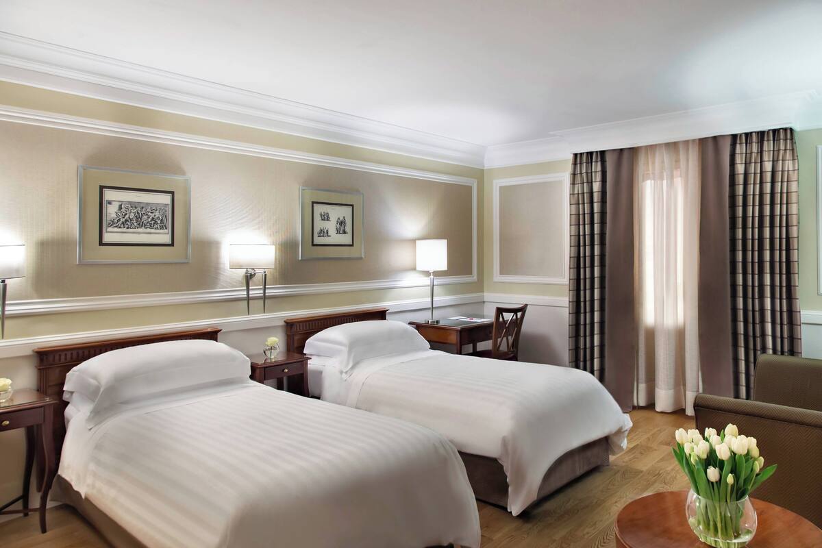 A hotel room with two beds, a desk, a chair, a lamp, and a vase of flowers.