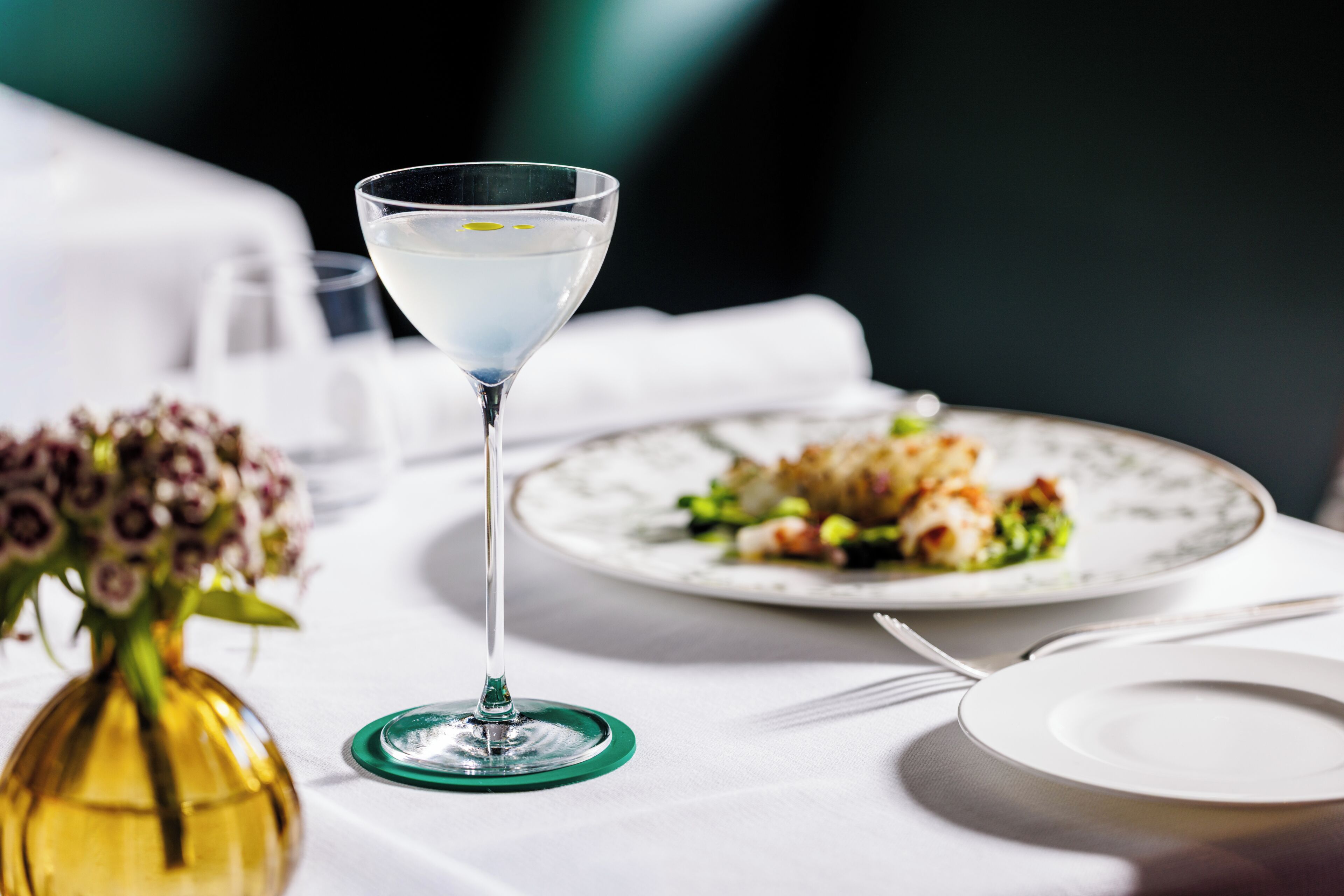 A dining table with a martini glass, a plate of food, and a vase with flowers.