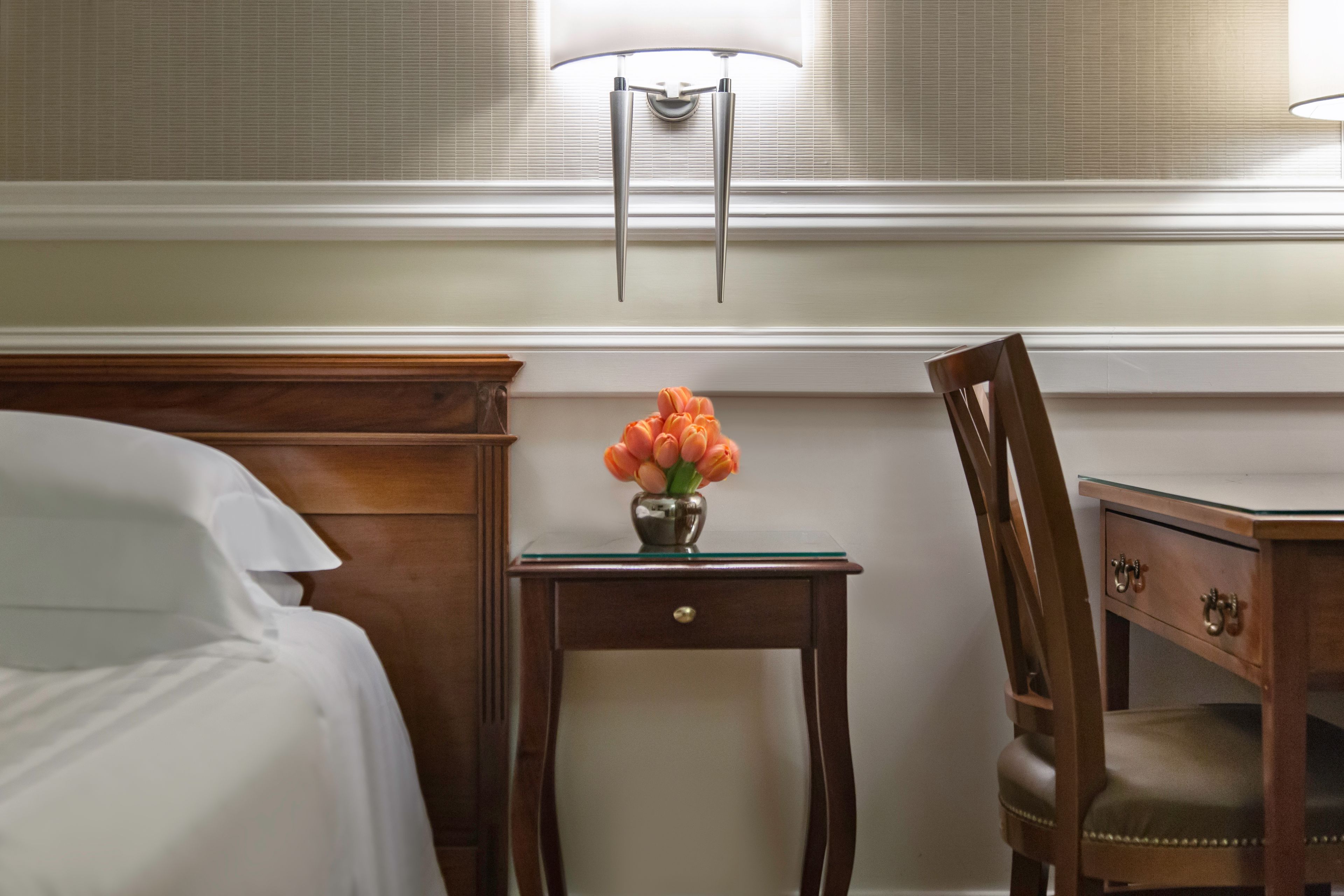 A neatly made bed with white linens, a wooden nightstand with a vase of orange flowers, a wooden chair, and a desk with drawers.