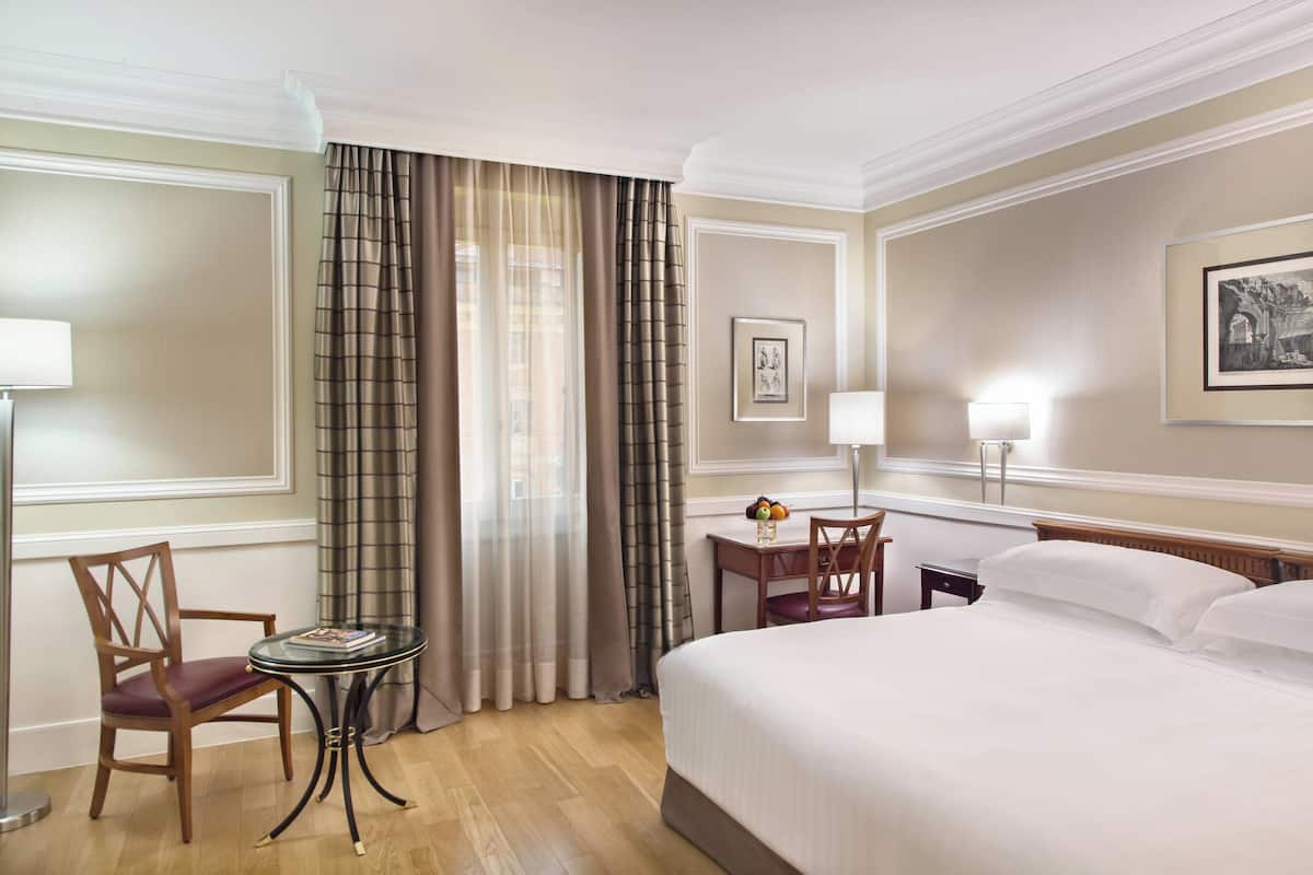 A hotel room with a bed, a chair, a small table with a lamp, and curtains.