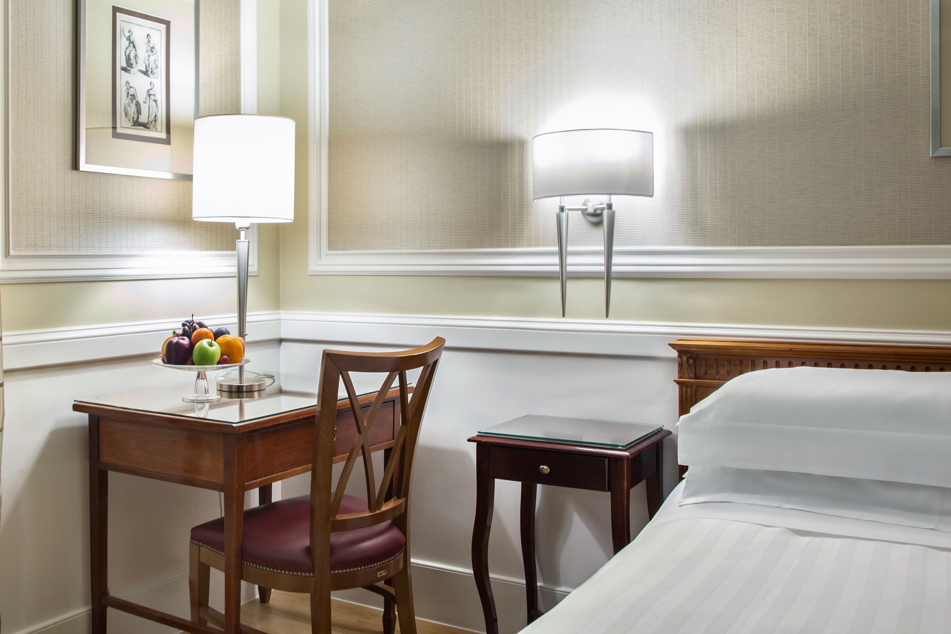 A hotel room with a bed, a desk with a lamp, a chair, a small table, and a framed picture on the wall.