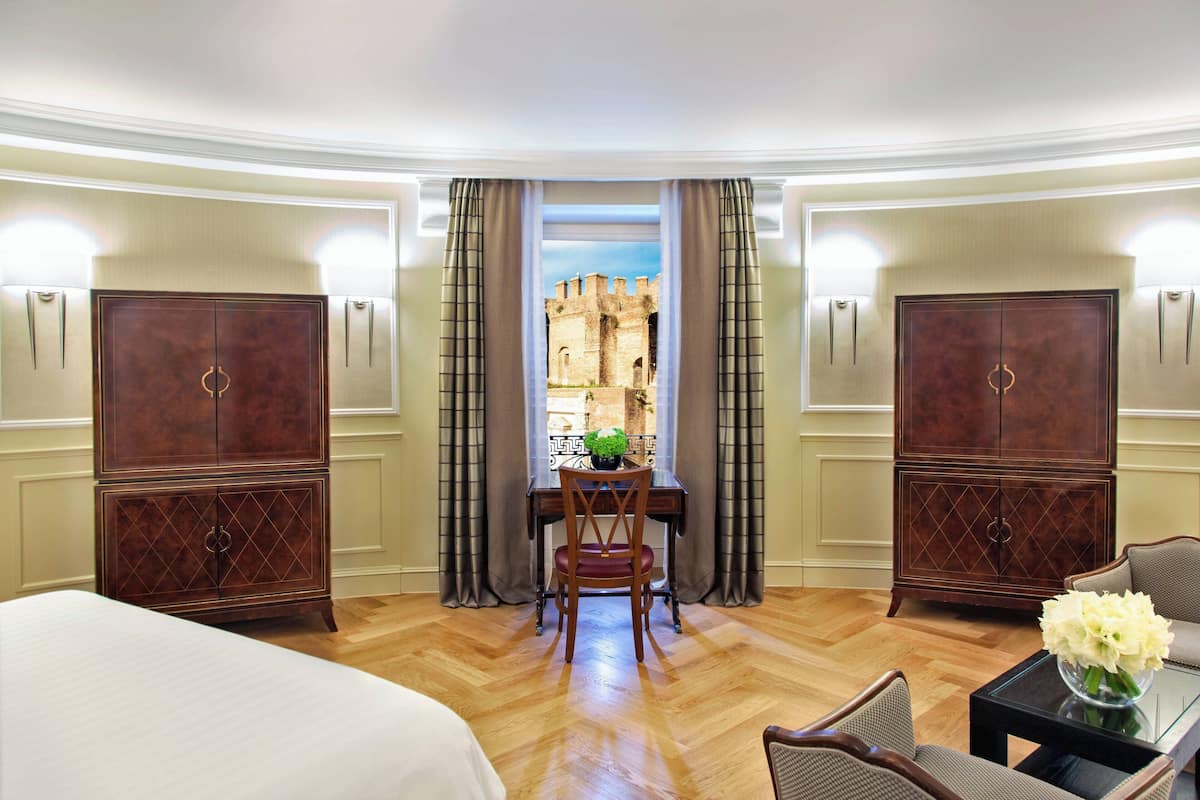 A hotel room with a large bed, a dining table with a chair, a small table with a vase, and two wardrobes.