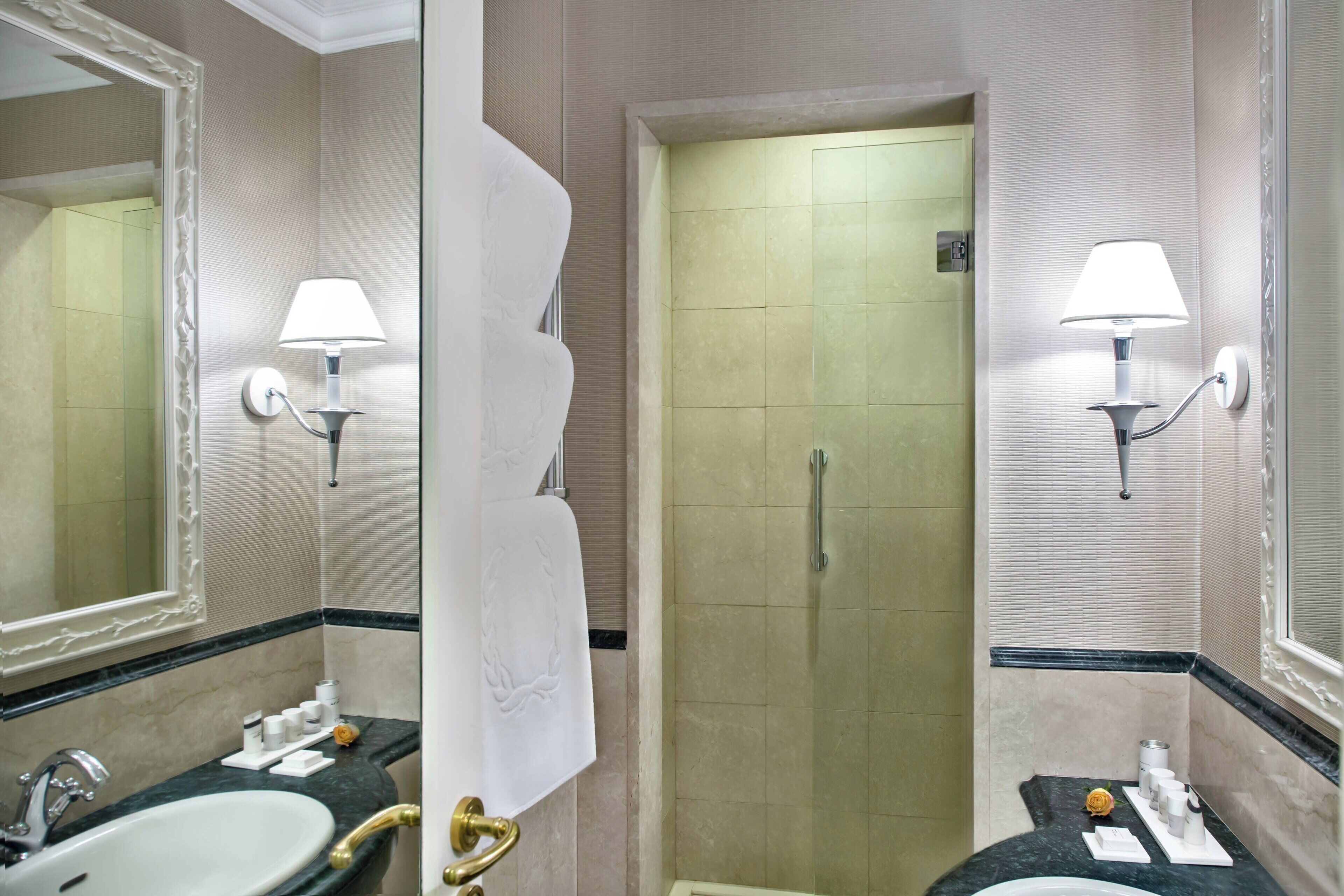 A bathroom with a sink, mirror, towel rack, shower, and two wall-mounted lamps.