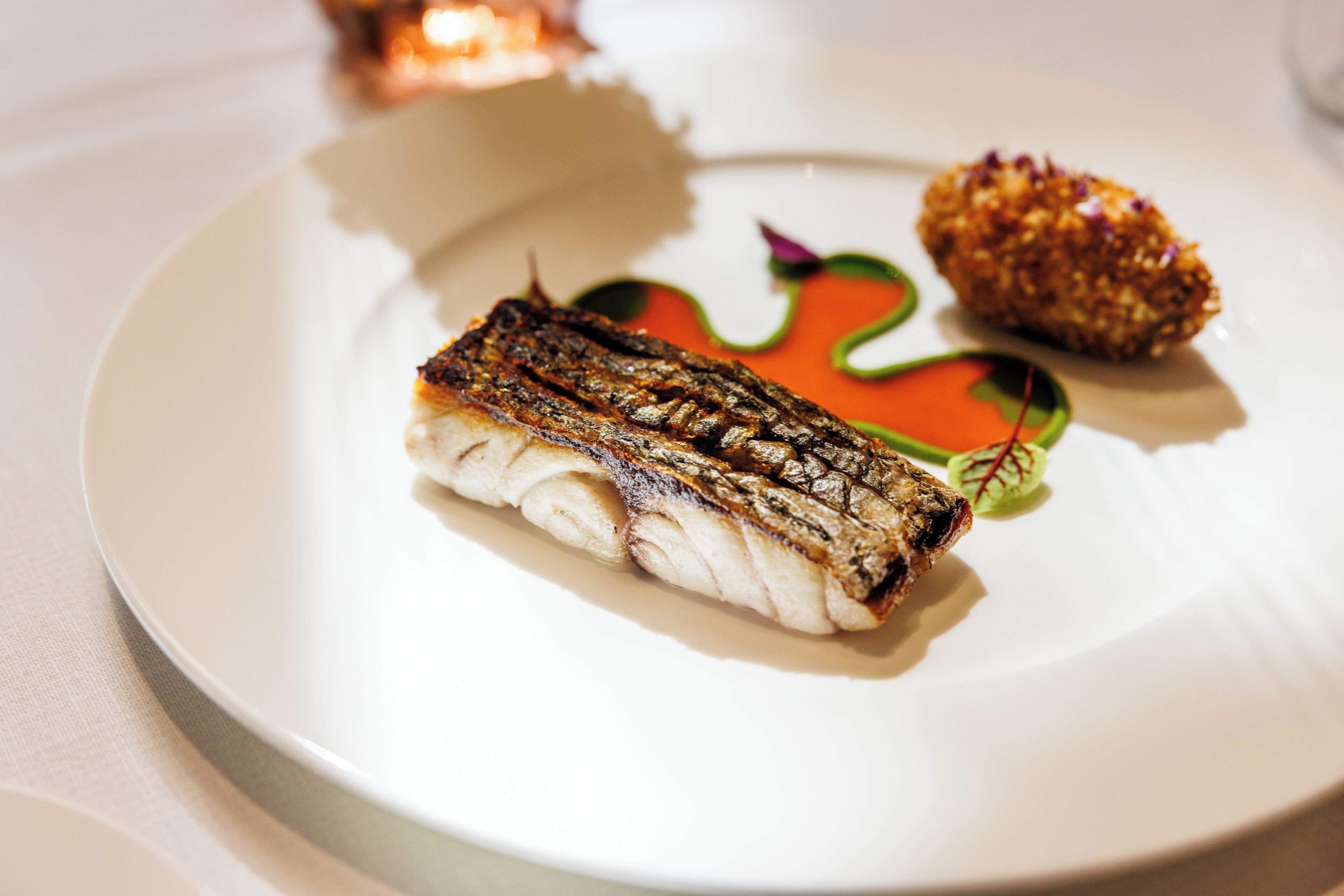 A plate with a piece of grilled fish, a small breaded item, and a colorful garnish.