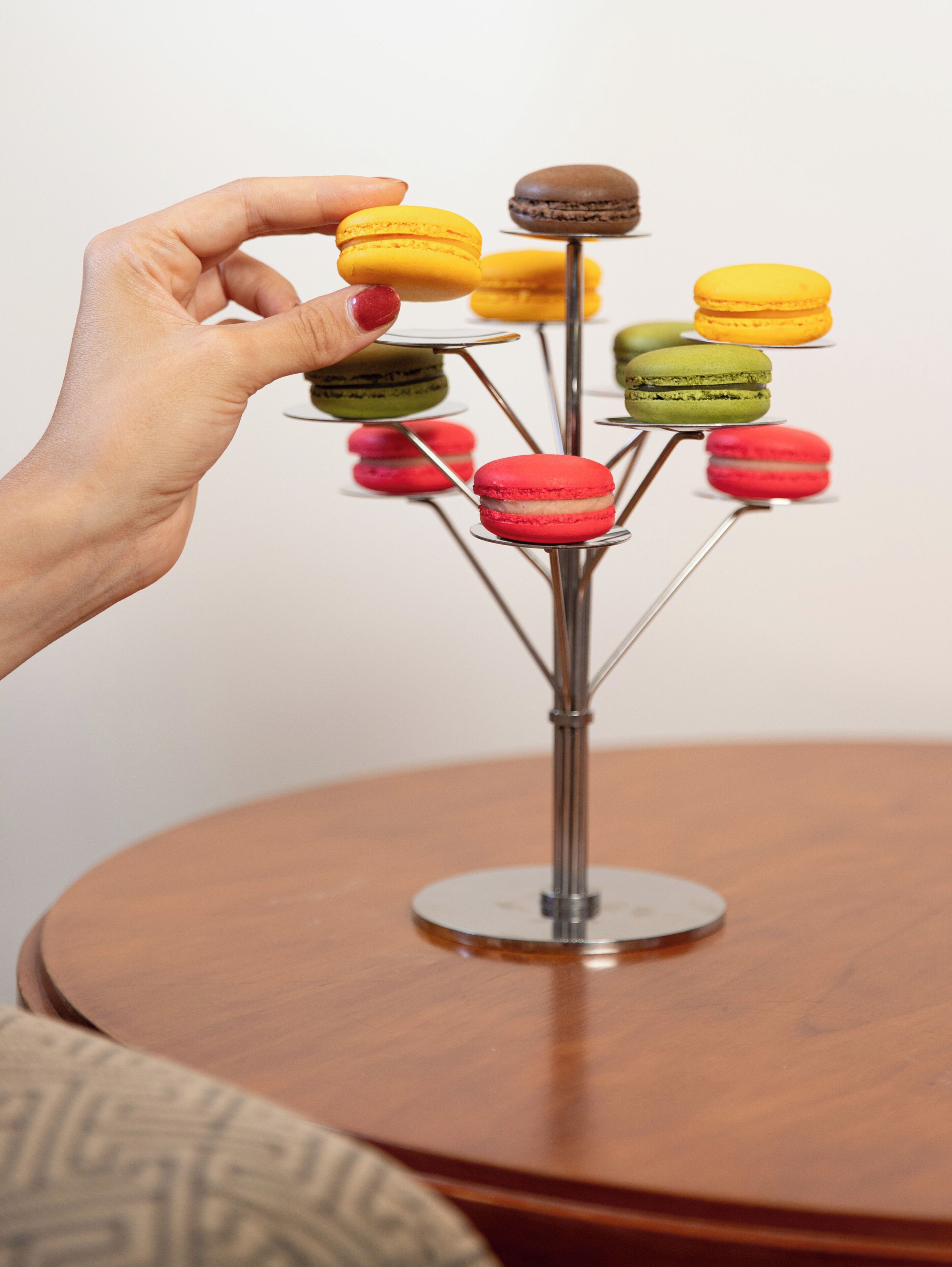 A hand holding a yellow macaron, with a macaron stand displaying various colored macarons.