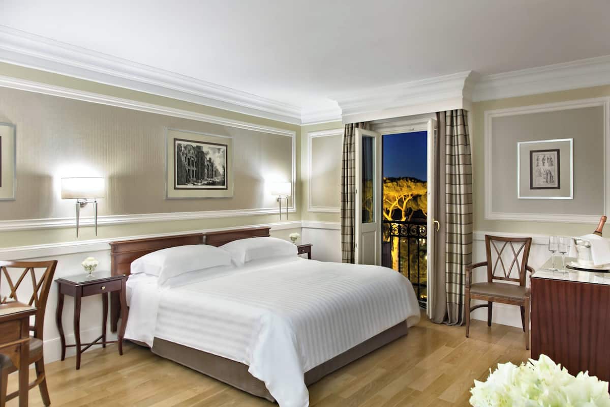 A hotel room with a large bed, a desk, a chair, and a view of a cityscape at night.