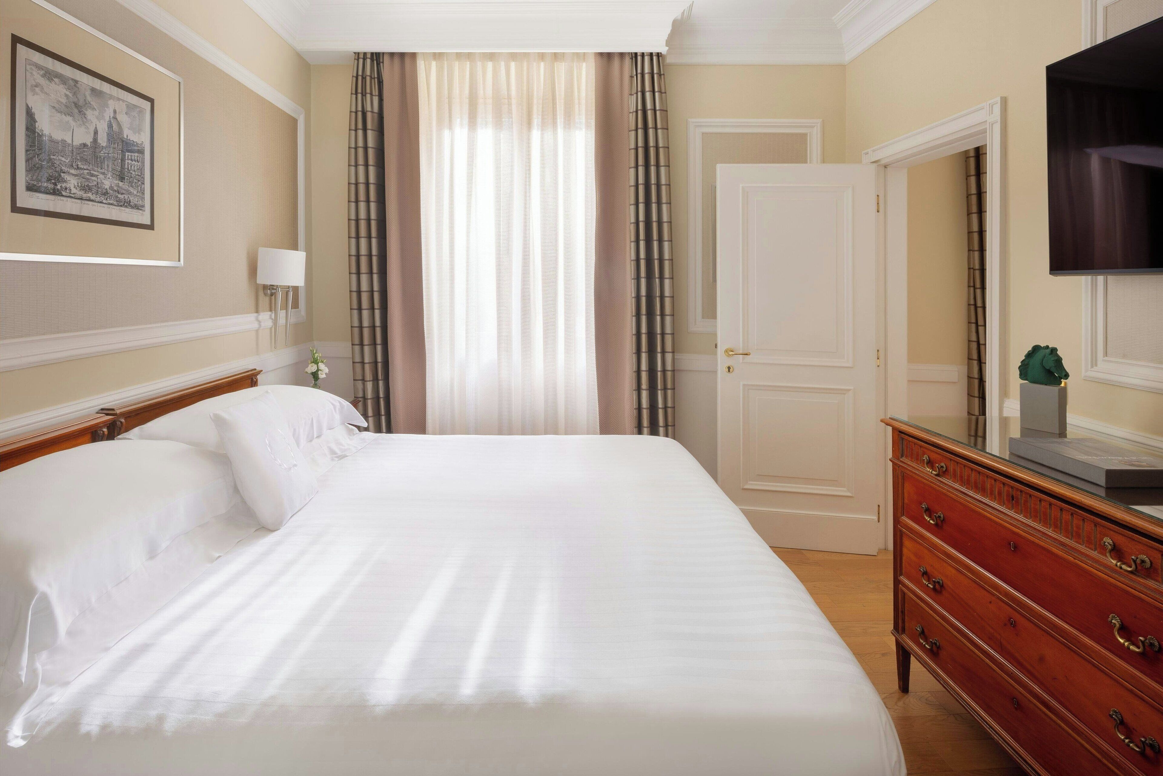 Una cama bien hecha con ropa blanca, un aparador de madera con cajones, un cuadro en la pared y una puerta con espejo.