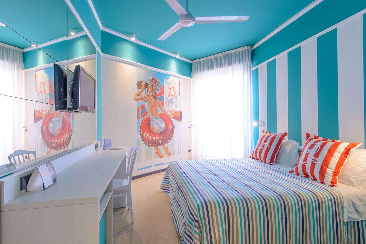 A room with a bed, a desk, a chair, a ceiling fan, and a wall with a mural.