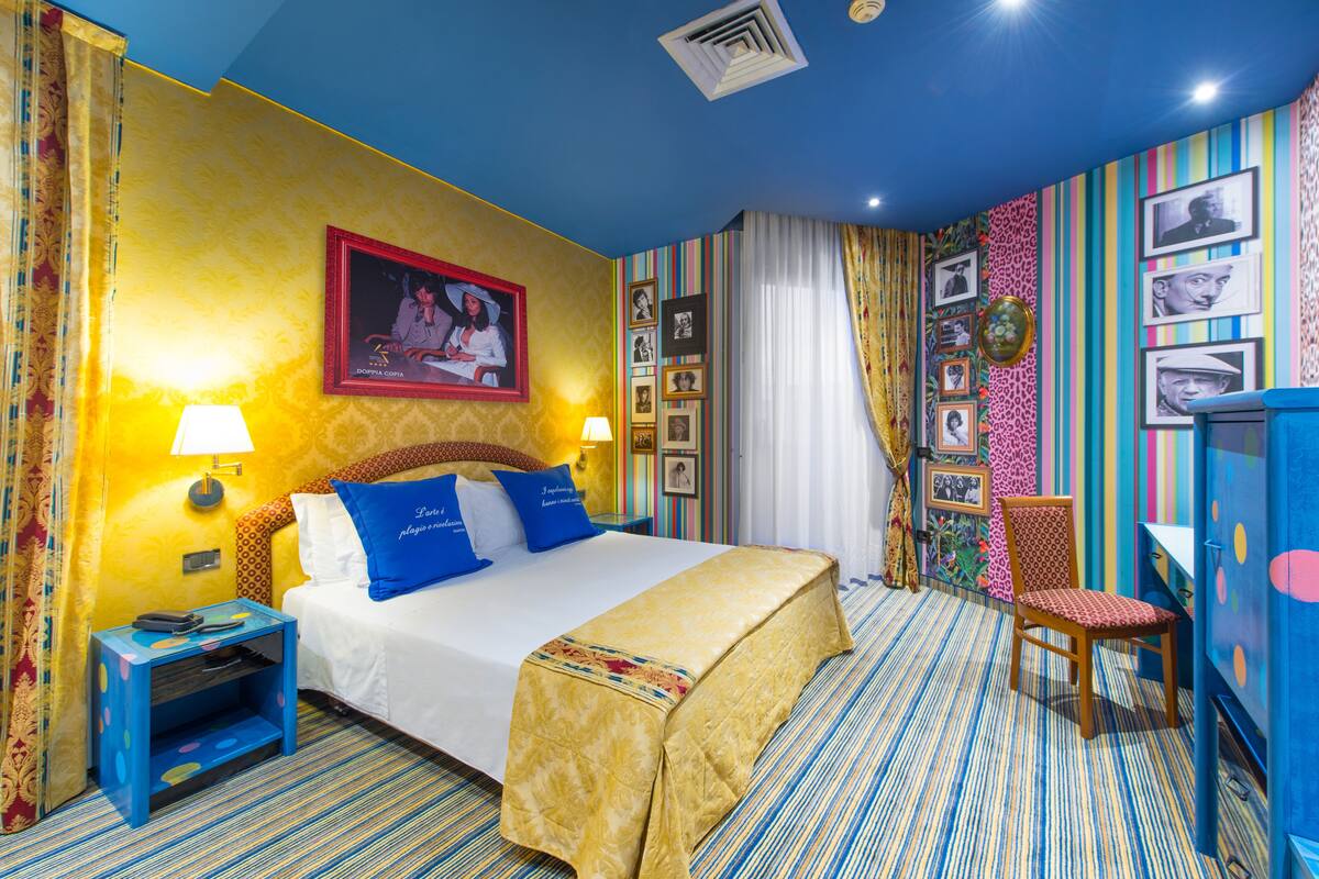 A hotel room with a bed, a chair, a desk, and a wall decorated with pictures.
