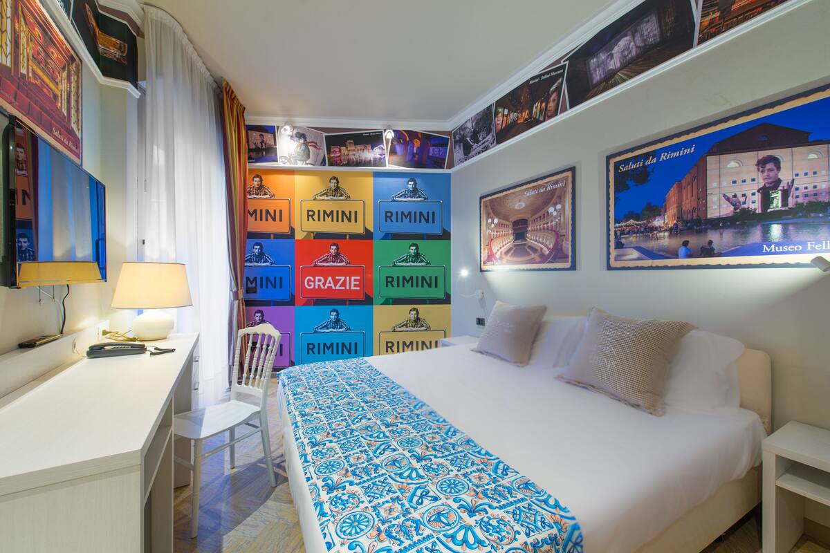 A hotel room with a bed, a desk, and a chair. The walls are decorated with colorful posters and pictures.