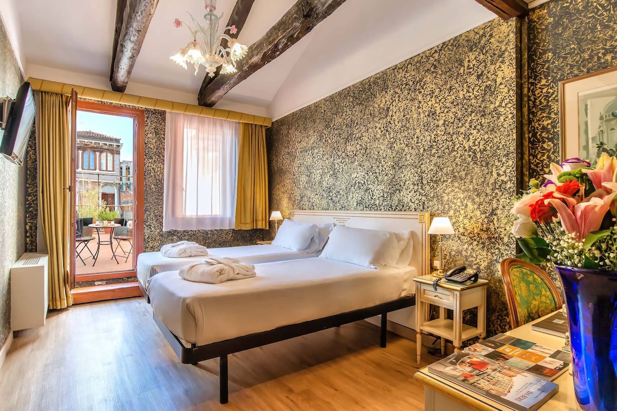 Une chambre d’hôtel avec un lit, un bureau, une chaise, un vase avec des fleurs et une vue sur l’extérieur.