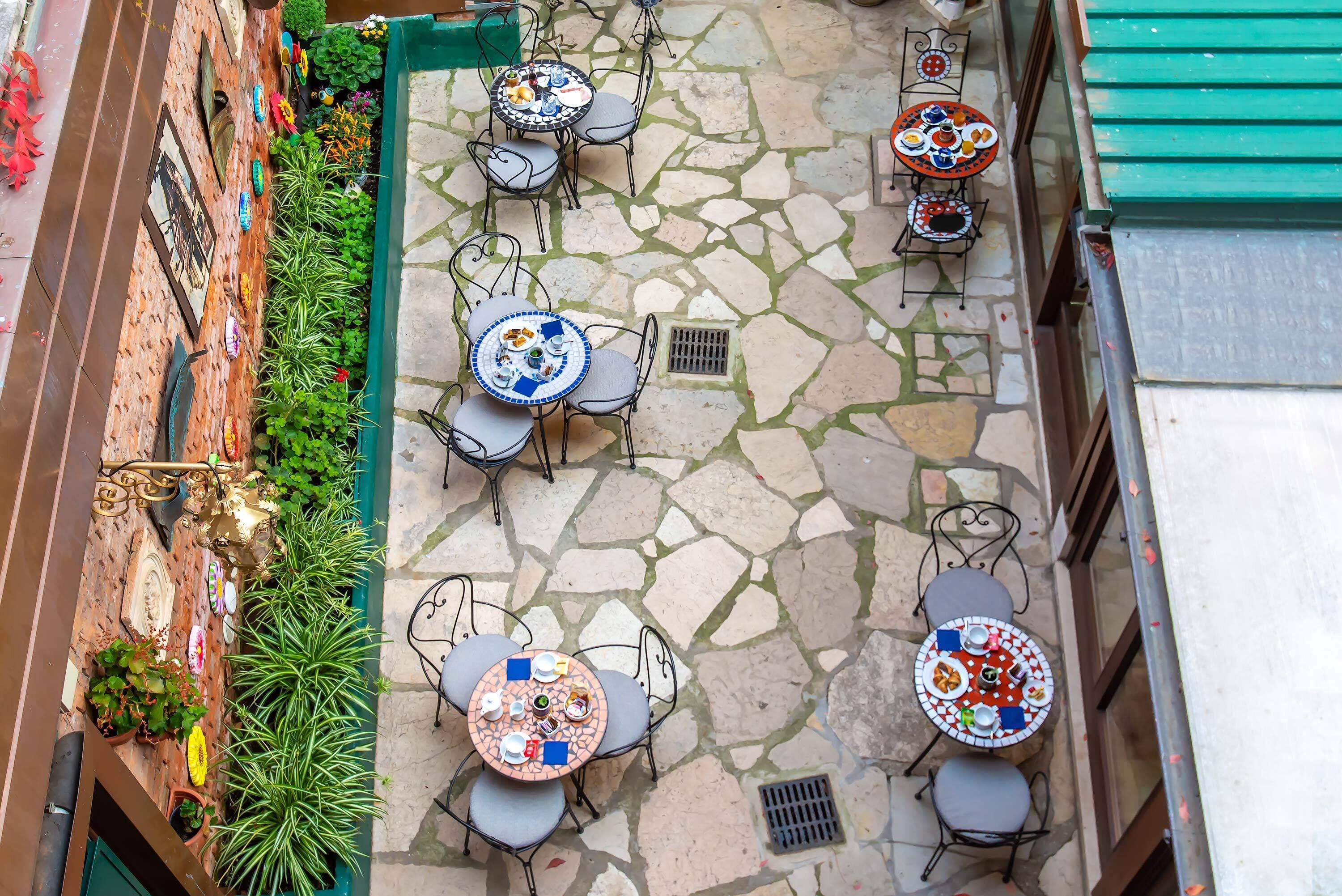 An outdoor patio area with stone flooring and multiple round tables with chairs, surrounded by potted plants and decorative items.