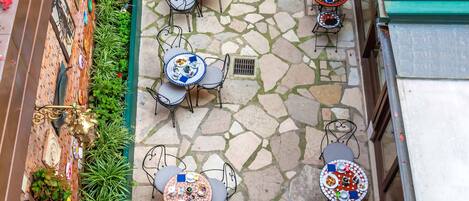 An outdoor patio area with stone flooring and multiple round tables with chairs, surrounded by potted plants and decorative items.