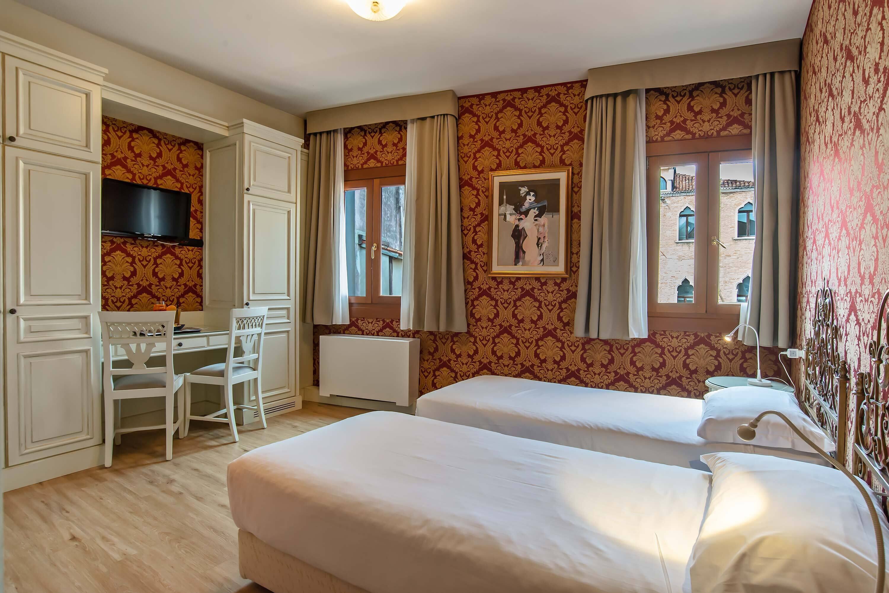 Quarto de hotel com duas camas, uma escrivaninha com cadeira, televisão e janela com cortinas.