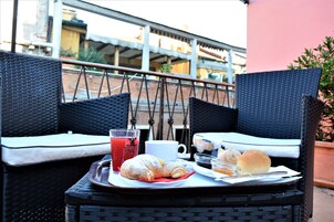 A wicker table with a tray containing food and drinks, two wicker chairs, and a view of buildings in the background.